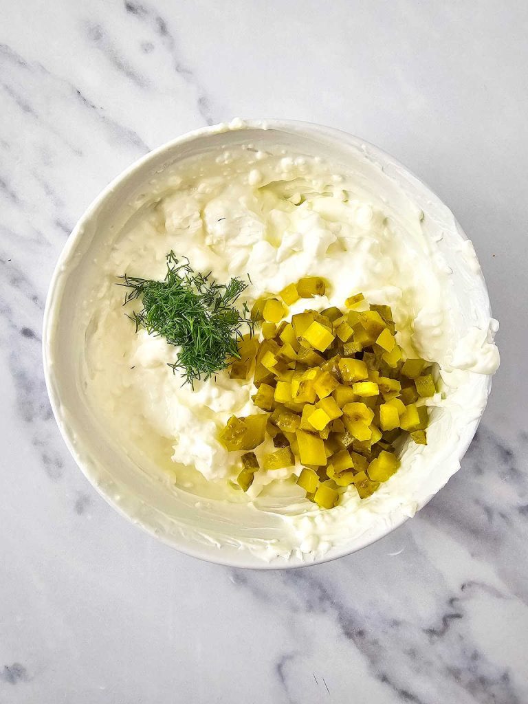 Chopped fresh dill and pickle on dip mixture in a mixing bowl.