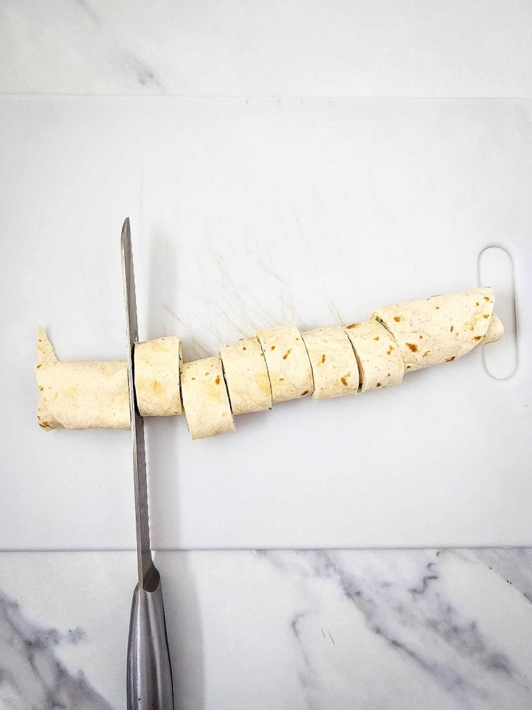 Knife cutting rolled up pickle dip pinwheel.