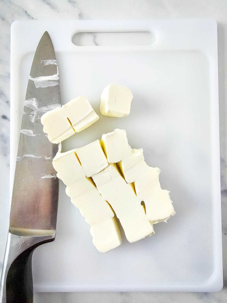 Small chunks of cut cream cheese and a knife on a white cutting board.