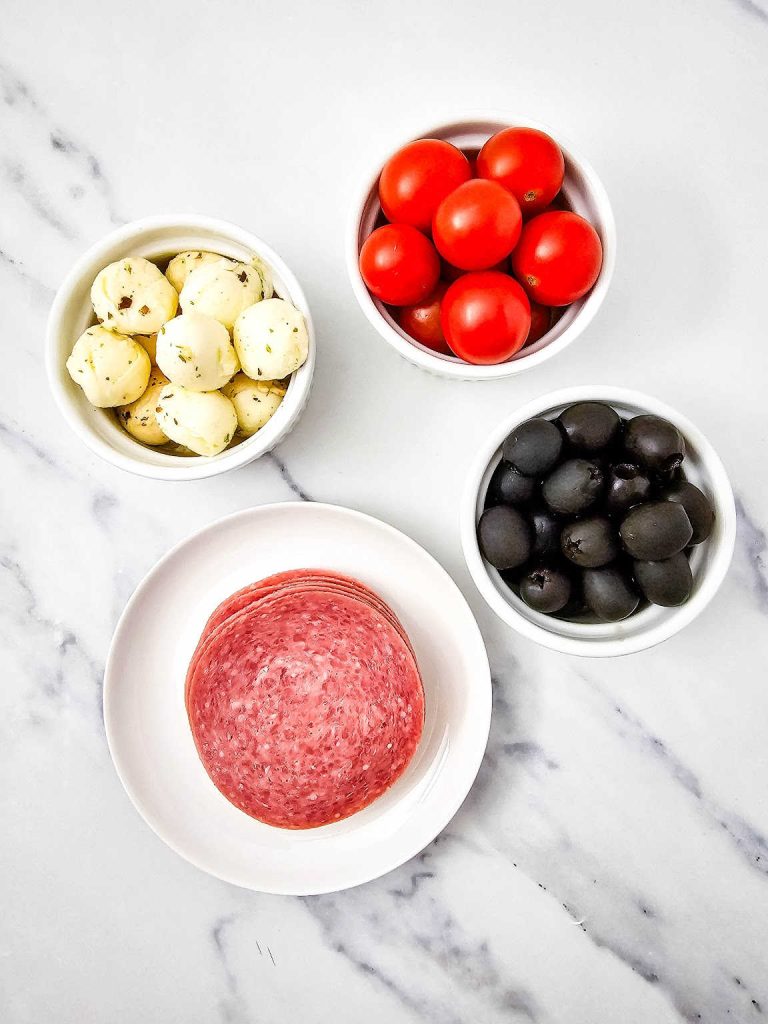 Olives, deli style hard salami, marinated mozzarella balls, cherry tomatoes on a marble countertop.