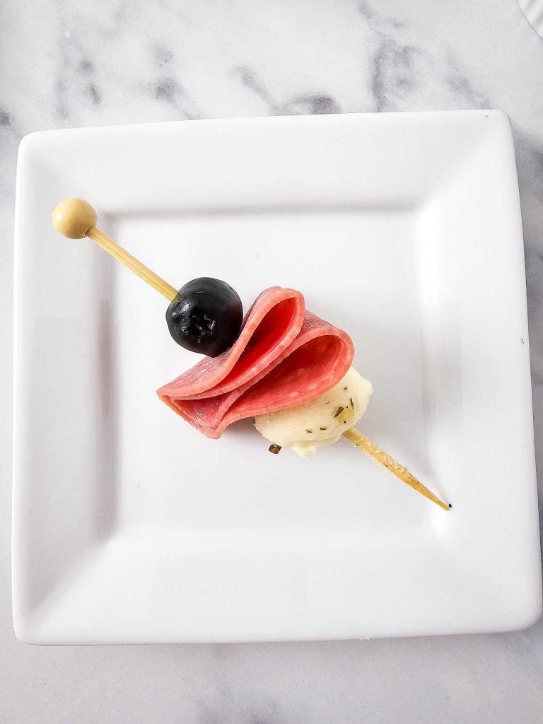 Skewered mozzarella ball, folded Salami and olive on a white plate.
