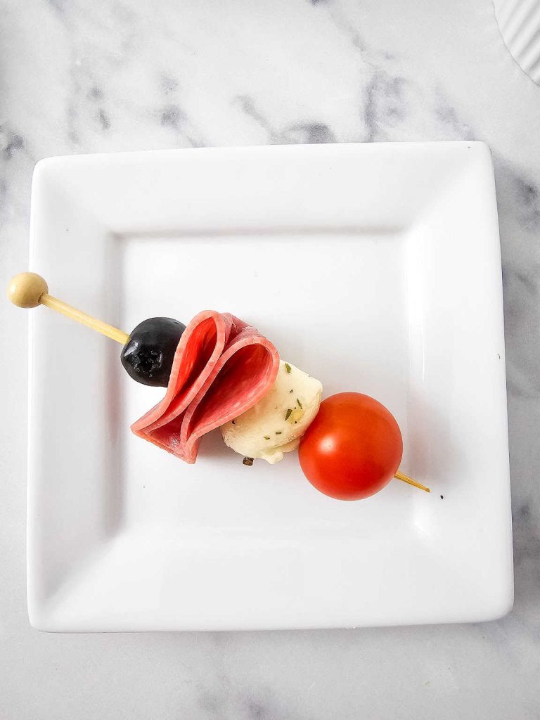 Skewered olive, folded Salami, mozzarella ball and cherry tomato on a white plate.