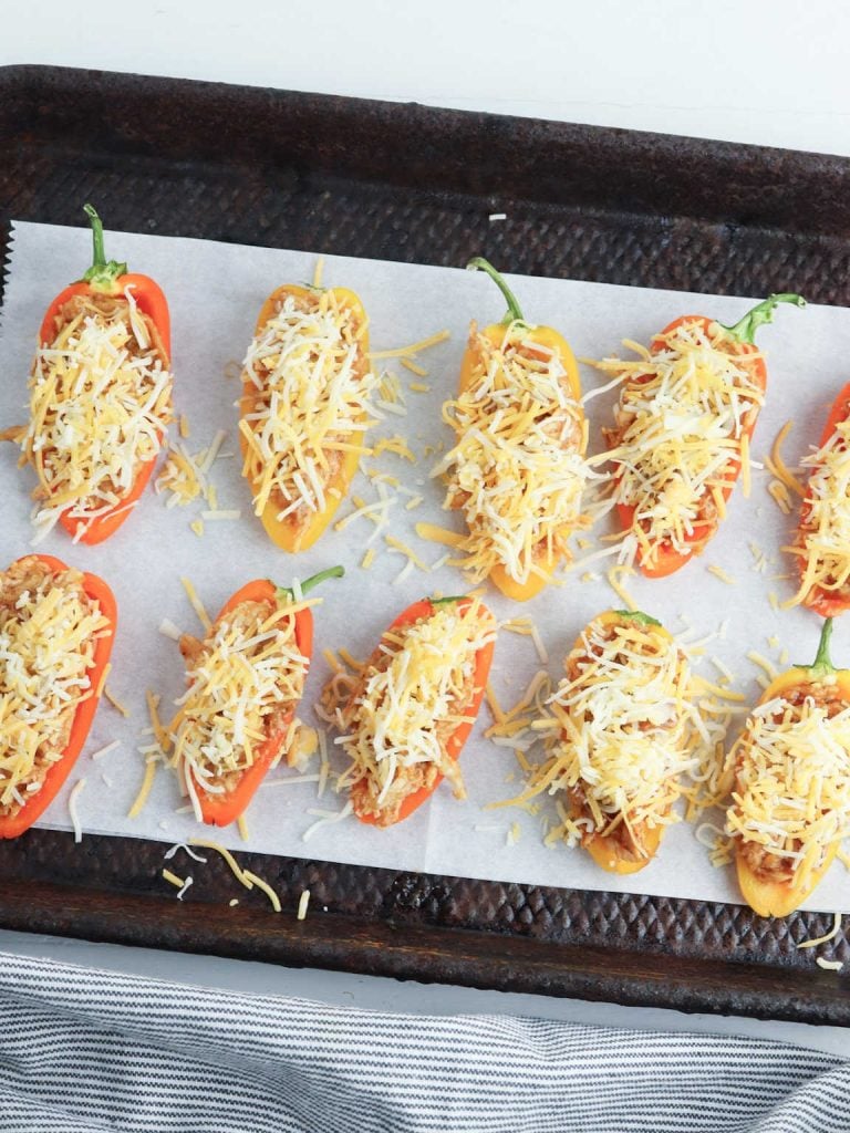 Mini peppers stuffed with shredded chicken mix and cheese on a parchment paper lined baking sheet.