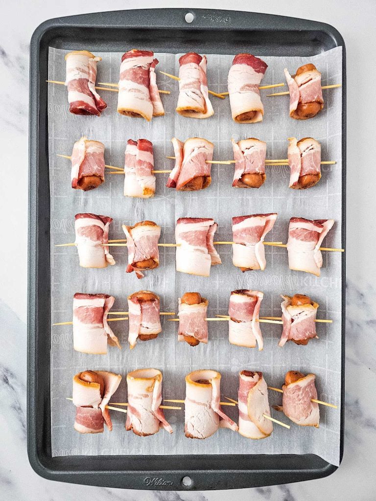 Uncooked bacon wrapped smokies on a parchment paper lined baking sheet.