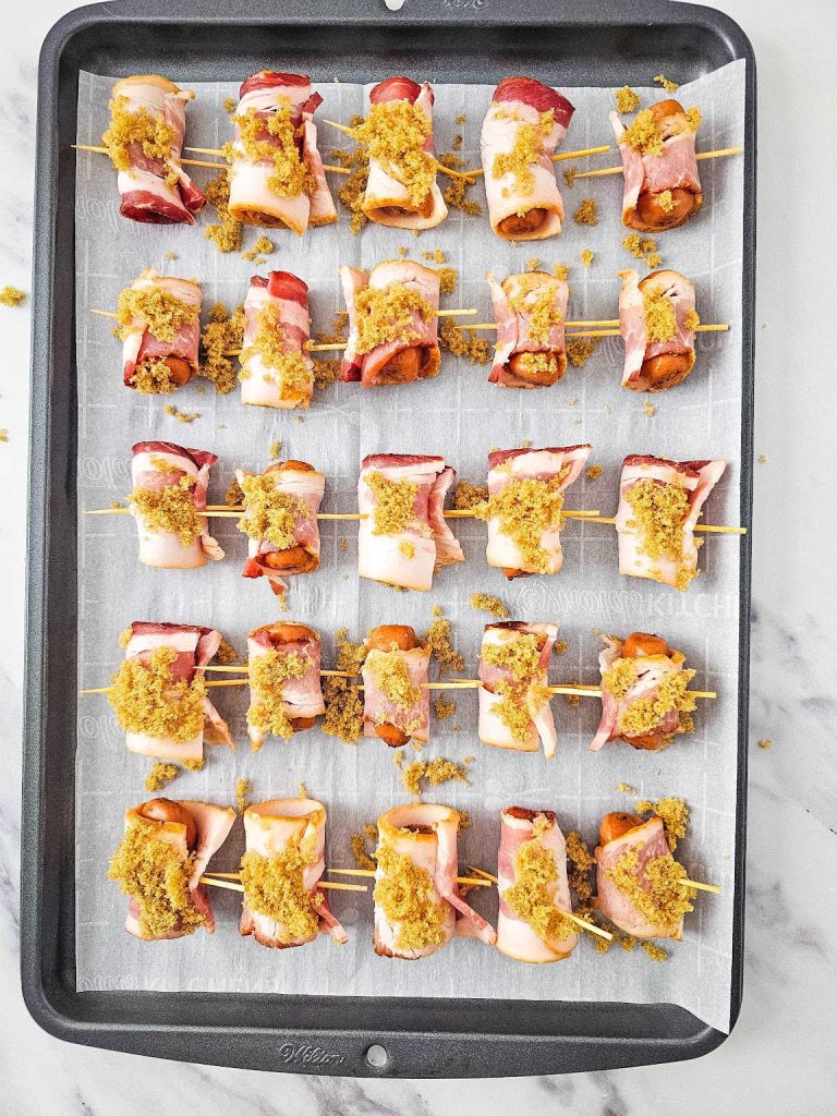 Uncooked bacon wrapped smokies sprinkled with brown sugar on a parchment paper lined baking sheet.
